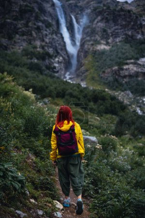 Foto de Una mujer, excursionista camina a lo largo de un sendero entre altas montañas. Cascada Shdugra en el fondo. Concepto de senderismo, estilo de vida activo, viajes. Día de verano, Georgia. Foto vertical - Imagen libre de derechos