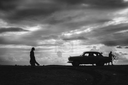 Téléchargez les photos : Silhouette d'un homme, femme avec véhicule rétro, debout sur la plage. Noir et blanc, nuages de fortes pluies grises. Coucher de soleil. Le concept de voyage et de loisirs sur les voitures classiques - en image libre de droit