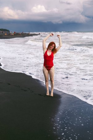 Photo for Happy woman tourist in red bodysuit. Standing on black sand, big stormy waves on the sea. Danger for swimming. Rain clouds on the horizon. Ureki Beach. Georgia. - Royalty Free Image