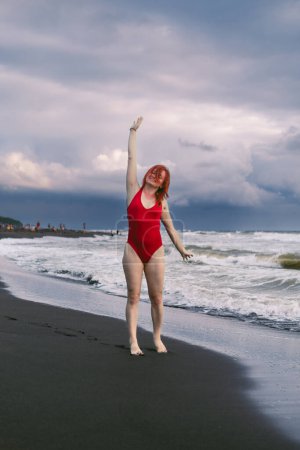 Photo for Happy woman tourist in red bodysuit. Standing on black sand, big stormy waves on the sea. Danger for swimming. Rain clouds on the horizon. Ureki Beach. Georgia. Vertical photo - Royalty Free Image