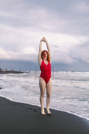 Photo for Happy woman tourist in red bodysuit. Standing on black sand, big stormy waves on the sea. Danger for swimming. Rain clouds on the horizon. - Royalty Free Image