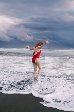 Photo for Happy woman tourist in red bodysuit. Standing on black sand, big stormy waves on the sea. Danger for swimming. Rain clouds on the horizon. feet in cold water. Vertical photo - Royalty Free Image