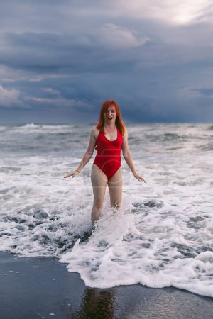 Photo for Happy woman tourist in red bodysuit. Standing on black sand, big stormy waves on the sea. Danger for swimming. Rain clouds on the horizon. feet in cold water. Vertical photo - Royalty Free Image