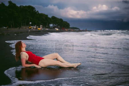 Photo for Happy woman tourist in red bodysuit. lies on the sand in the water, big stormy waves on the sea. Danger for swimming. Rain clouds on the horizon. - Royalty Free Image