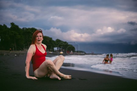 Photo for Happy woman tourist in red bodysuit. Standing on black sand, big stormy waves on the sea. Danger for swimming. Rain clouds on the horizon. - Royalty Free Image