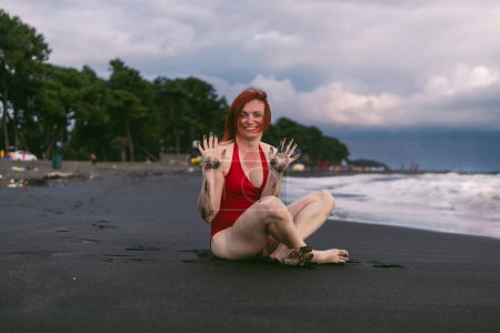 Photo for Happy woman tourist in red bodysuit shows dirty hands. Sitting on the black sand near the water, waves on the sea. Rain clouds on the horizon. - Royalty Free Image
