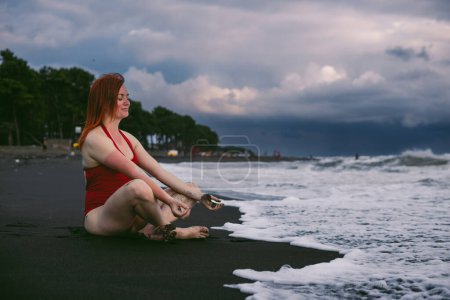 Photo for Happy woman tourist in red bodysuit Sitting in the position, yoga on black sand, stormy waves on the sea. Danger for swimming. Rain clouds on the horizon. Ureki besch, Georgia - Royalty Free Image