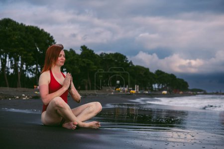Photo for Happy woman tourist in red bodysuit Sitting in the lotus position, yoga on black sand, stormy waves on the sea. Danger for swimming. Rain clouds on the horizon. Ureki besch, Georgia - Royalty Free Image