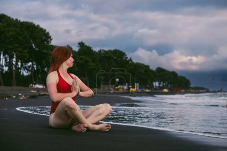 Photo for Happy woman tourist in red bodysuit Sitting in the lotus position, yoga on black sand, stormy waves on the sea. Danger for swimming. Rain clouds on the horizon. Copy space. Ureki besch, Georgia - Royalty Free Image