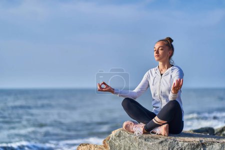 Photo for Serene tranquil one woman in lotus position meditates and relaxes on a cliff against the background of the sea and clear blue sky. Calm concept and balance of mental health - Royalty Free Image