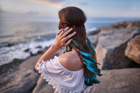 Téléchargez les photos : Hippie indie femme avec des plumes bleues dans les cheveux sur le bord de mer au coucher du soleil. Ambiances Boho et mode bohème - en image libre de droit