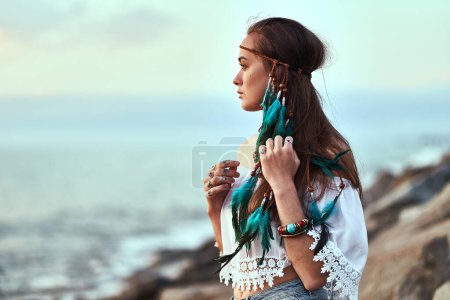 Foto de Retrato de mujer hippie atractiva con plumas azules en el pelo largo, joyas y blusa blanca en la orilla del mar. Estilo boho chic - Imagen libre de derechos