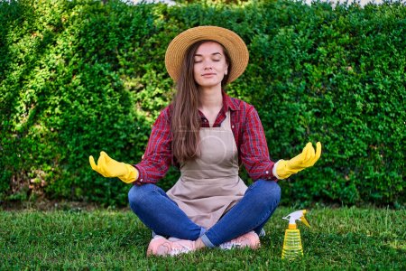 Photo for Calm woman gardener in a straw hat, apron and yellow rubber gloves in lotus posture meditates and relaxes on green grass in the flowers garden. Mental health and enjoying of gardening - Royalty Free Image