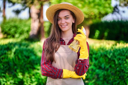 Foto de Retrato de feliz alegre sonrisa atractiva mujer jardinero en sombrero de paja, delantal y guantes de goma amarilla celebración botella de aerosol para regar las plantas en el jardín en el día soleado - Imagen libre de derechos