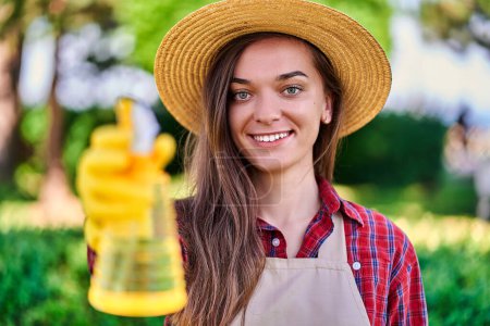 Téléchargez les photos : Portrait de heureuse femme souriante et joyeuse jardinière en chapeau de paille avec vaporisateur jaune pour arroser les plantes dans le jardin par temps ensoleillé - en image libre de droit