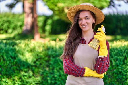 Foto de Retrato de feliz mujer atractiva jardinero sonriente en sombrero de paja, delantal y guantes de goma amarilla con botella de spray para regar plantas en jardín verde en día soleado - Imagen libre de derechos