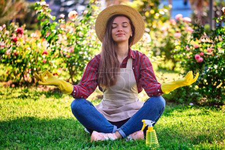 Téléchargez les photos : Femme calme jardinier dans un chapeau de paille, tablier et gants en caoutchouc jaune en position lotus médite et se détend sur l'herbe dans le jardin de fleurs. Santé mentale et plaisir du jardinage - en image libre de droit