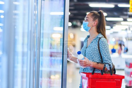 Foto de Mujer joven comprador casual en una máscara de protección médica con cesta de la compra elige un productos lácteos del congelador en una tienda de comestibles - Imagen libre de derechos