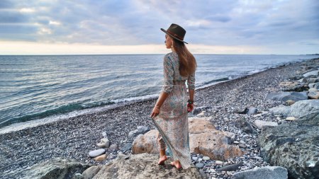 Téléchargez les photos : Femme Boho en robe longue et chapeau en feutre debout sur la pierre au bord de la mer - en image libre de droit
