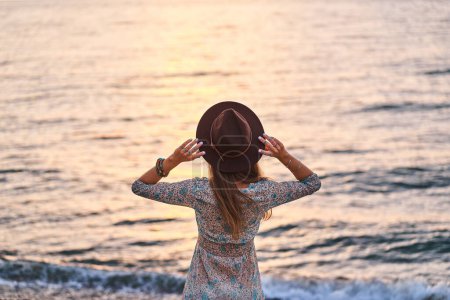 Téléchargez les photos : Femme boho sereine libre portant une robe et un chapeau en feutre debout au bord de la mer au coucher du soleil et profitant du calme et de la liberté - en image libre de droit