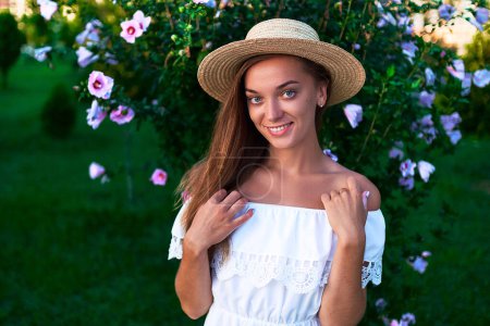 Foto de Retrato de joven feliz mujer romántica sonriente con el pelo largo en un vestido blanco de verano con hombros desnudos y un sombrero de paja en un parque de flores en verano - Imagen libre de derechos