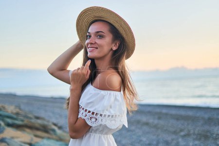 Foto de Retrato de hermosa joven feliz mujer romántica sonriente en un vestido blanco de verano con hombros desnudos y un sombrero de paja a la orilla del mar en verano - Imagen libre de derechos