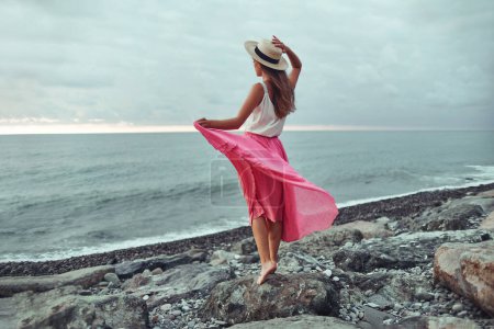 Foto de Mujer romántica soñando en blusa blanca, falda larga rosa ondeando y sombrero de paja de pie en la orilla del mar y mirando a la distancia - Imagen libre de derechos