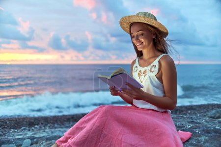 Foto de Sonriendo feliz soñando mujer romántica leyendo libro de novela en la orilla del mar y disfrutando de un momento feliz en la vida - Imagen libre de derechos