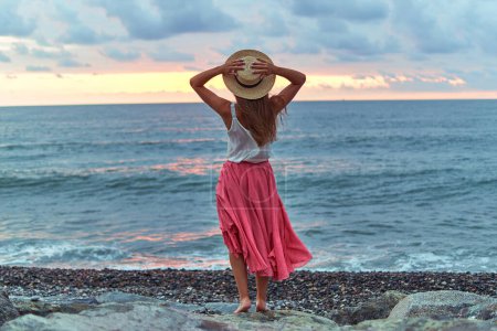 Foto de Mujer romántica soñando en blusa blanca, falda larga rosa y sombrero de paja de pie en la orilla del mar y disfrutando de un momento feliz en la vida y una libertad - Imagen libre de derechos
