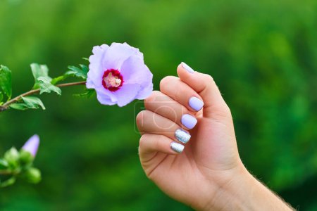 Foto de Manicura femenina con esmalte de uñas de color y brillo plateado sobre el fondo de las flores en un parque al aire libre - Imagen libre de derechos