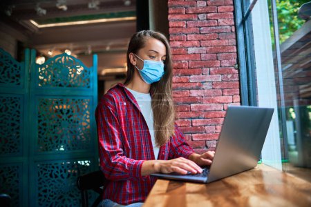 Foto de Una joven freelancer que usa una mascarilla de medicina trabajando remotamente en una computadora en un café durante una pandemia. Distancia social y protección sanitaria frente a virus en lugares públicos - Imagen libre de derechos