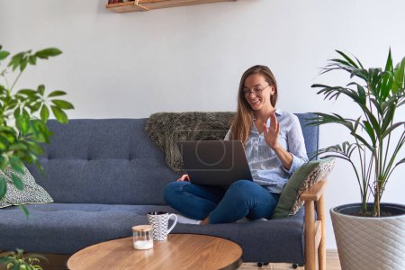 Photo for Young casual happy smiling woman in round glasses sitting on the sofa and communicate virtually with family and friends on a laptop during comfy cozy home rest among indoors plants - Royalty Free Image
