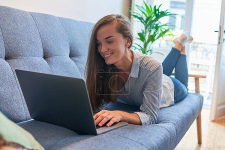 Foto de Feliz sonrisa joven mujer casual acostada en un sofá, descansando y navegando en línea en un ordenador portátil en el acogedor hogar cómodo - Imagen libre de derechos
