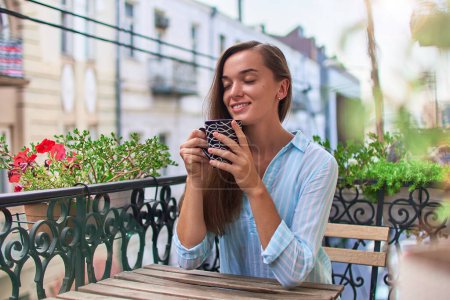 Foto de Hermosa mujer romántica feliz sonriente con los ojos cerrados disfrutando de café aromático en el balcón por la mañana temprano en la ciudad - Imagen libre de derechos