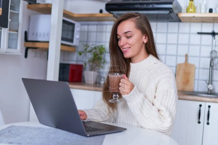 Photo for Happy smiling cozy woman wearing a warm white sweater drinking a hot cocoa and browsing online at laptop at comfy homey kitchen - Royalty Free Image