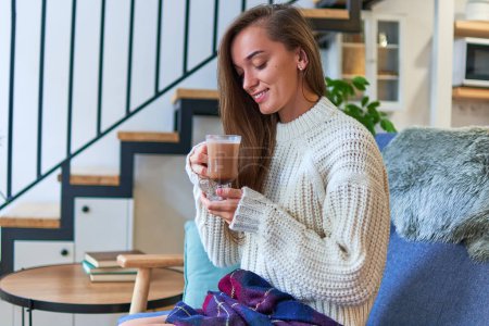 Téléchargez les photos : Portrait d'une jolie jeune femme souriante heureuse et confortable dans un pull chaud blanc buvant du cacao chaud à la maison confortable - en image libre de droit