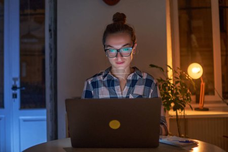 Photo for Concentrated woman workaholic in glasses works online in the evening at a laptop at home - Royalty Free Image