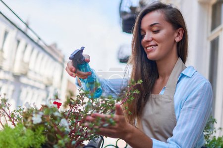 Téléchargez les photos : Joyeux sourire femme mignonne jardinier portant tablier arrosage balcon fleurs à l'aide de vaporisateur - en image libre de droit