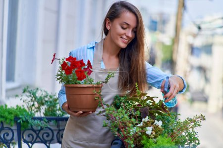 Happy smiling cute woman gardener wearing apron holding flower pot petunia and taking care about balcony plants  