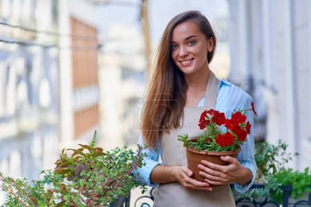 Photo for Portrait of happy smiling attractive woman gardener wearing apron holding flower pot on a balcony - Royalty Free Image