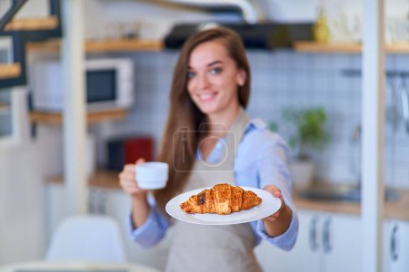 Téléchargez les photos : Portrait d'une belle femme joyeuse et joyeuse chef pâtissier souriant avec une assiette de croissant brun doux frais cuit au four et tasse de café - en image libre de droit