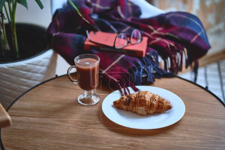 Photo for A blanket, book, fresh crispy croissant on a plate and a cup of hot cocoa on a wooden table for a comfort homely pastime, rest and relaxation at cozy atmosphere - Royalty Free Image