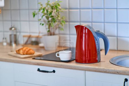 Foto de Hervidor eléctrico rojo moderno con una taza blanca en la mesa en la cocina en casa - Imagen libre de derechos