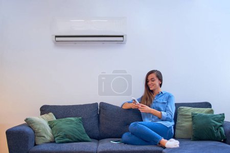 Foto de Mujer feliz joven sentada en el sofá bajo el aire acondicionado y ajustando la temperatura de confort con control remoto en el hogar moderno - Imagen libre de derechos