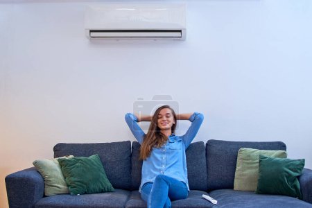 Photo for Young cute casual calm woman holding hands behind head with closed eyes resting on the comfortable couch under the air conditioner at modern home - Royalty Free Image
