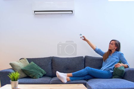 Foto de Mujer feliz joven sentada en el sofá bajo el aire acondicionado y ajustando la temperatura de confort con control remoto en el hogar moderno - Imagen libre de derechos
