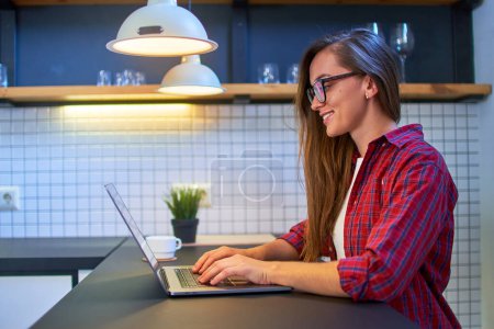 Téléchargez les photos : Jeune femme intelligente décontractée et souriante heureuse dans des lunettes et une chemise à carreaux travaillant à distance en ligne à l'ordinateur portable à la maison loft moderne - en image libre de droit