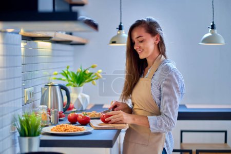 Photo for Smiling happy joyful cute cooking woman housewife cutting a fresh organic ripe tomatoes on chopping board for preparation dinner at modern kitchen - Royalty Free Image