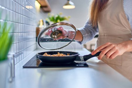 Photo for Woman preparing food in a frying pan on the stove for dinner at modern loft style kitchen - Royalty Free Image