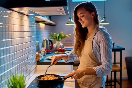 Téléchargez les photos : Joyeux sourire attrayant jeune cuisinière femme au foyer préparant la nourriture dans une poêle sur la cuisinière pour le dîner du soir à la cuisine moderne de style loft - en image libre de droit